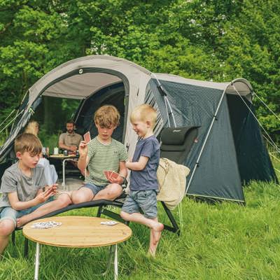 På billedet ses en familie på campingtur i et telt fra Outwell. De voksne slapper af i teltet, mens børnene spiller kort udenfor på et campingbord.