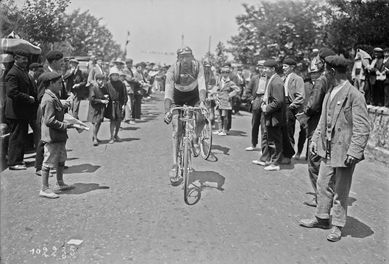 Billedet viser en cykelrytter der cykler på ruten, mens fans står og kigger på.