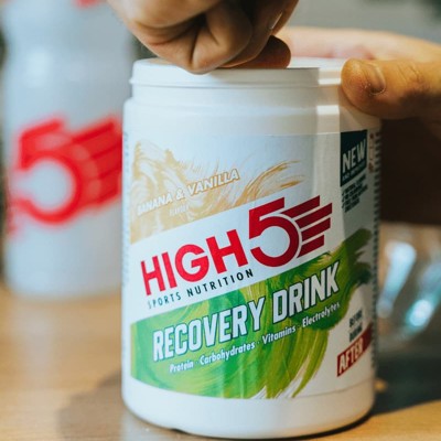 Billedet viser en bøtte med High5 recovery drink. På billedet kan man se en hånd som er ved at tage en skefuld pulver fra bøtten.