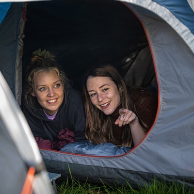 Billedet viser to kvinder der ligger og kigger ud af et festivaltelt. Den ene peger på noget og det er tydeligt at de hygger sig og har en god samtale..